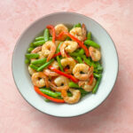 a finished dish of stir-fried garlic shrimp with snap peas and red pepper in a ceramic bowl on a pink background