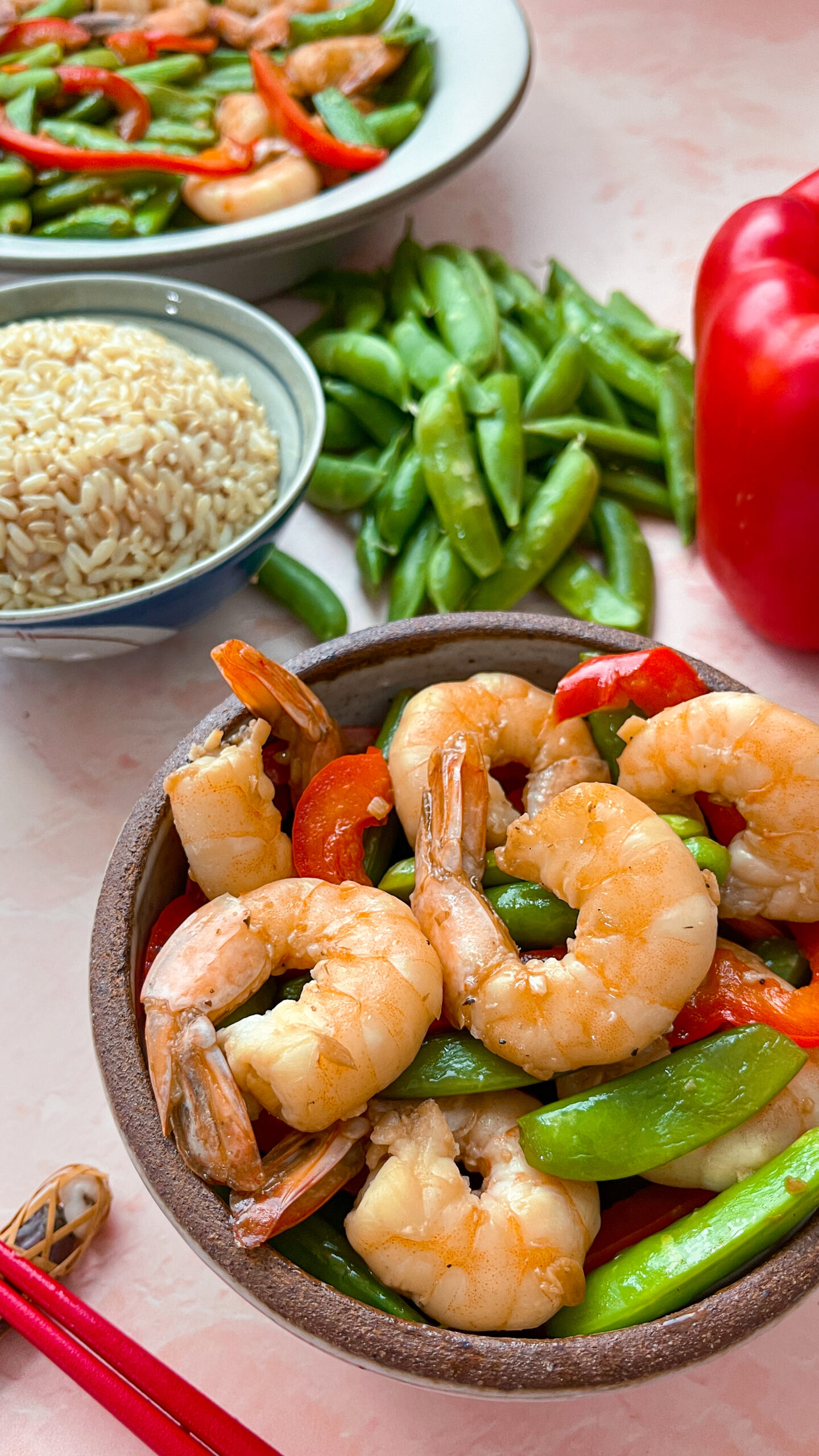 stir-fry shrimp with snap peas surrounded by a bowl of brown rice, and a large serving bowl of the dish