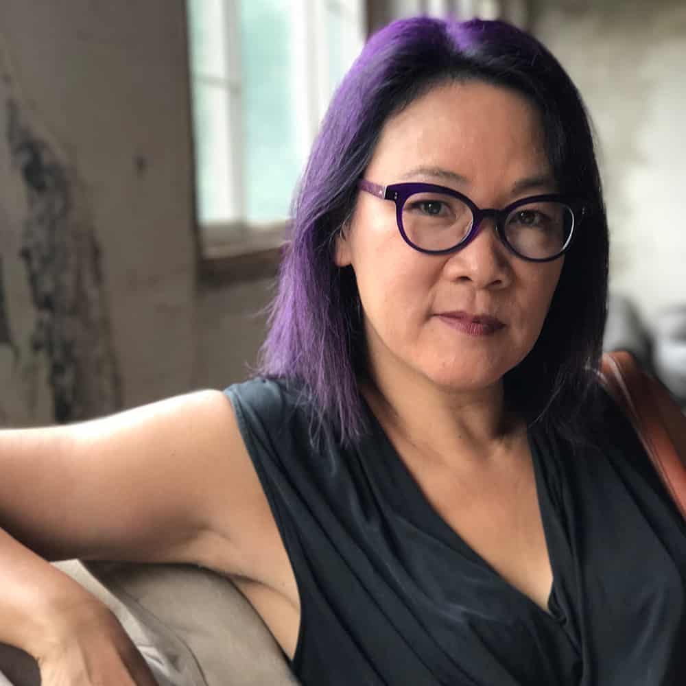 Photo of Carole Yu Food Blogger, She has purple hair, purple glasses a v neck black silky shirt on sitting on a couch with a window in the background