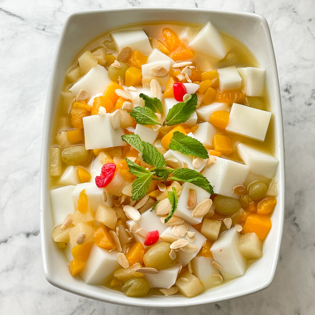 Close up of large bowl of Chinese Almond Jelly mixed in with fruit cocktail, mandarin oranges and topped with mint leaves