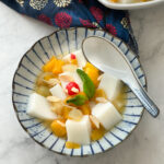 Easy Chinese Almond Jelly in a beautiful Japanese white bowl with blue ripple pattern. Almond Jelly is white cubes of jelly, surrounded by fruit cocktail, mandarin oranges, and mint.