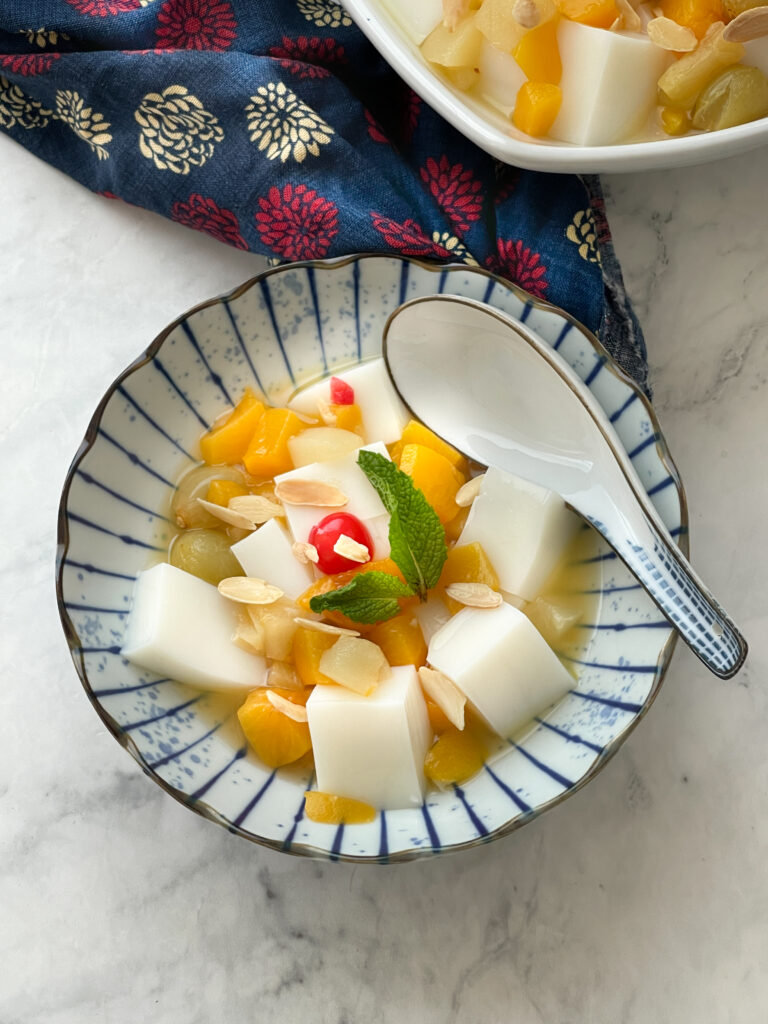 Easy Chinese Almond Jelly in a beautiful Japanese white bowl with blue ripple pattern. Almond Jelly is white cubes of jelly, surrounded by fruit cocktail, mandarin oranges, and mint.