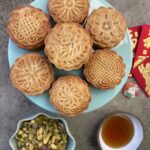 overhead shot of golden baked chinese honey pistachio mooncakes, with pistachios, and honey in accompanying bowls, with chinese red packets, and a rabbit button next to the cakes