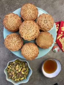 overhead shot of golden baked chinese honey pistachio mooncakes, with pistachios, and honey in accompanying bowls, with chinese red packets, and a rabbit button next to the cakes