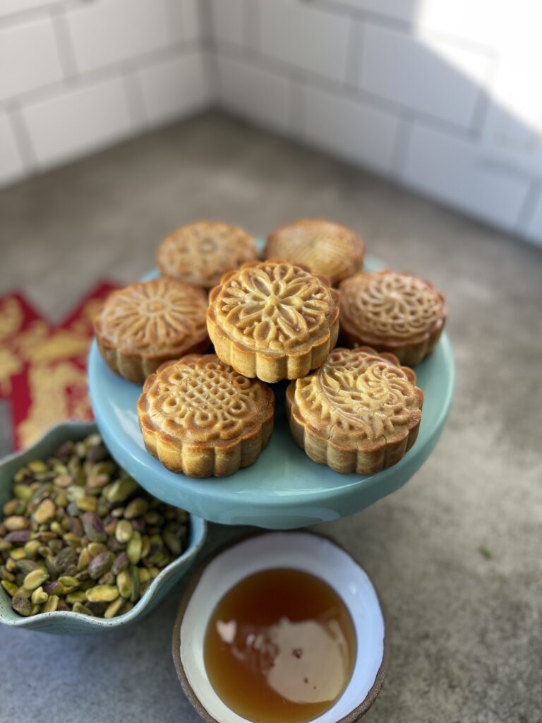 upclose of seven gorgeous honey pistachio mooncakes shiny and golden sitting on a small turquoise cake plate, with chinese money packets, a bowl of pistachios and a dish of honey on the concrete counter below