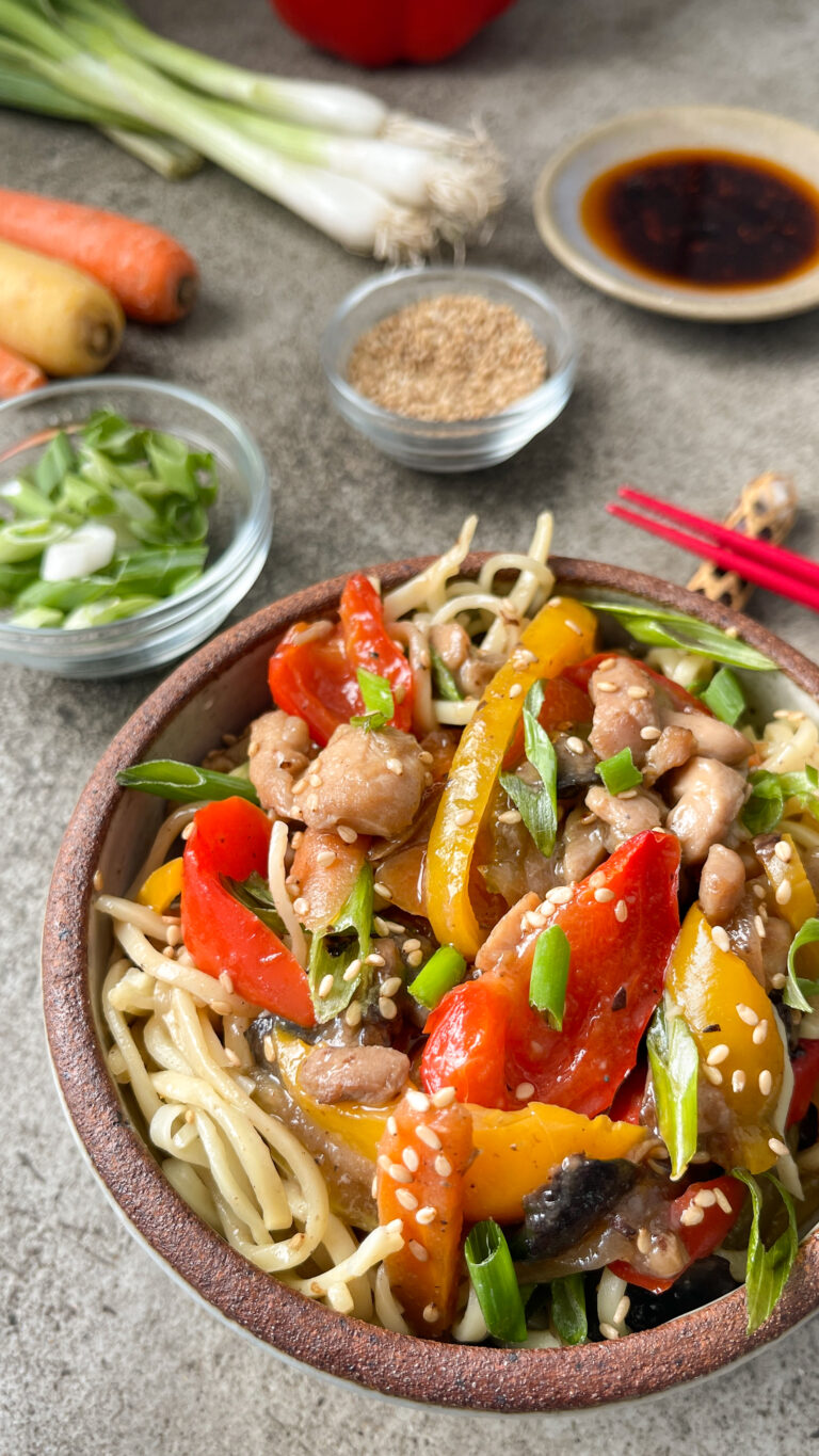 Bowl of Chicken Chow Mein noodles with red and yellow peppers, chicken, topped with green onions and sesame seeds