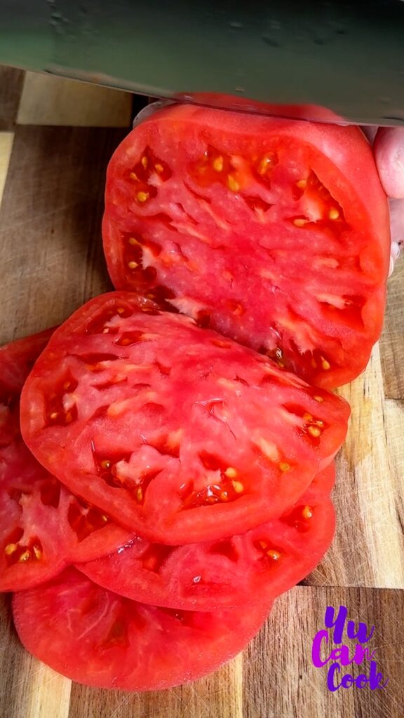 A juicy red heirloom tomato being cut into 1/4" slices on a bamboo cutting board.