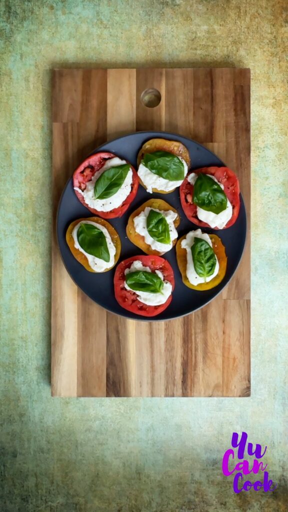 A Dark gray plate topped with slices of juicy red heirloom tomatoes topped with buffalo mozzarella, and a vibrant green leaf of basil on each slice