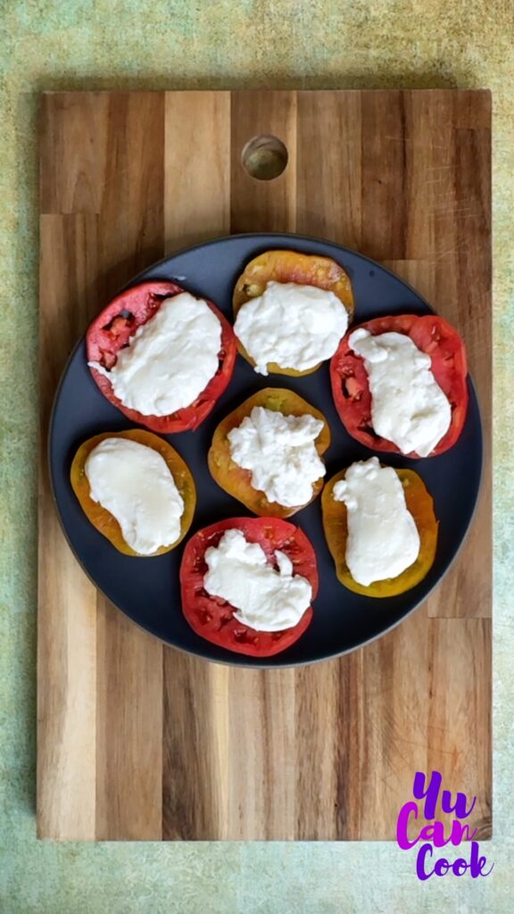 Dark Grey platter with juicy slices of red heirloom tomatoes, topped with buffalo mozzarella, all on a bamboo cutting board on top of a green mottled photography board.